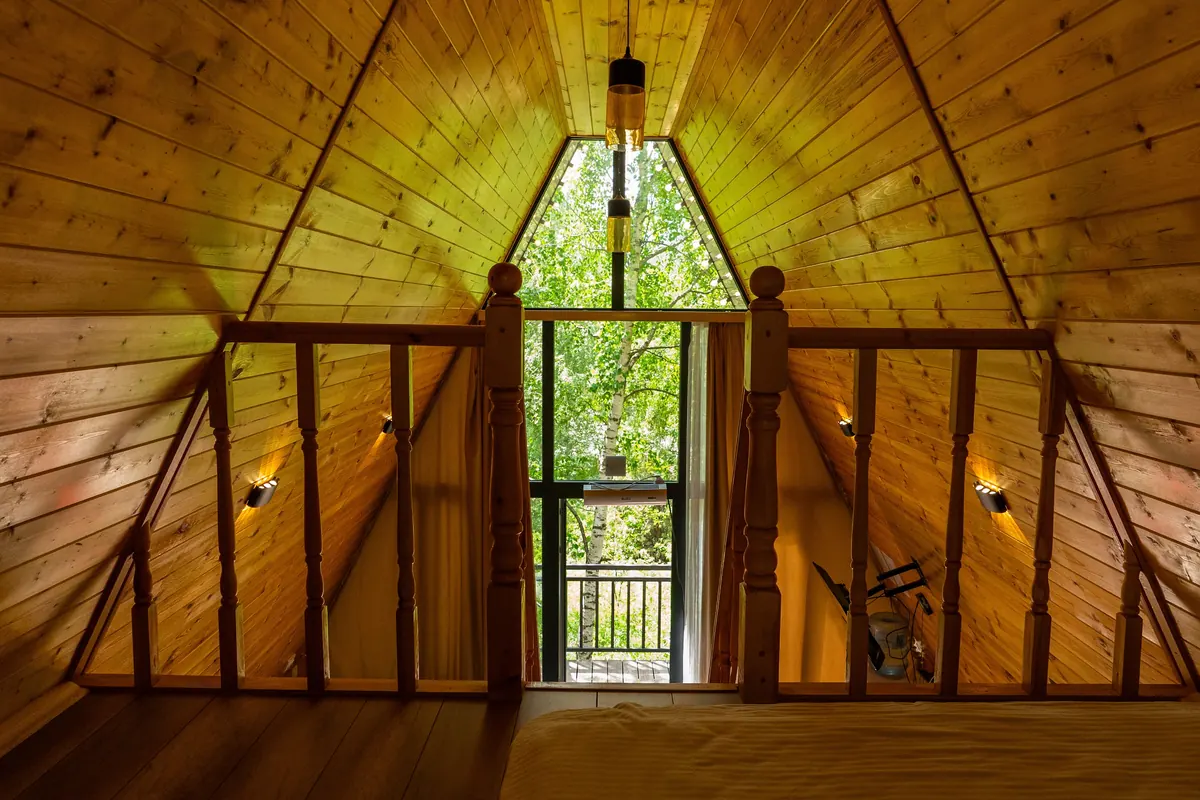 View from the A-frame loft: pine-clad sloping walls, a wooden balustrade and a tall triangular window onto green trees.